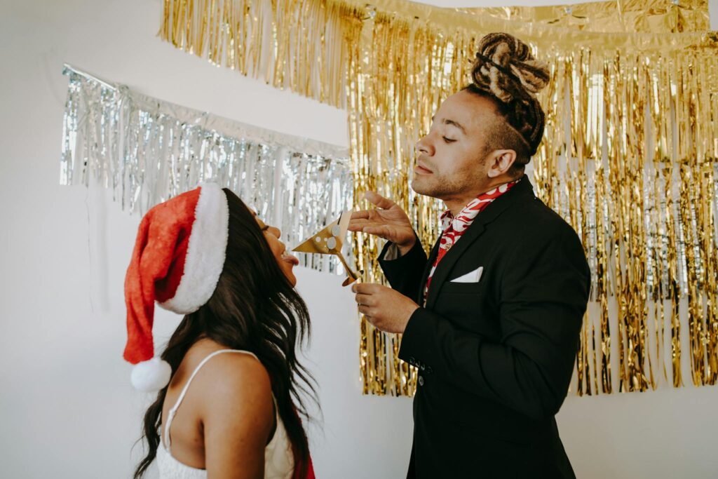Two adults enjoying a festive Christmas party with golden decorations and a Santa hat indoors.